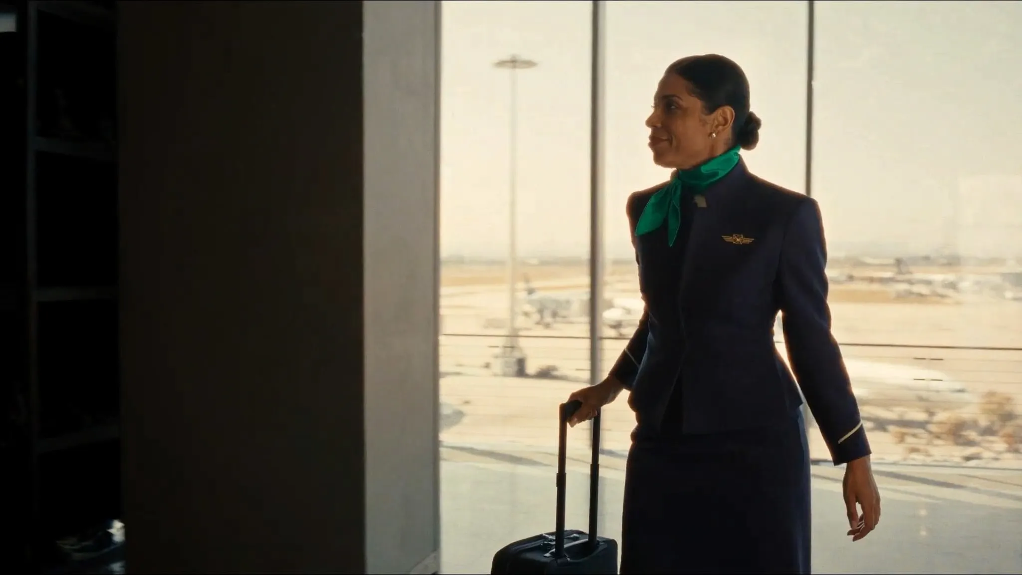 Comissária de voo de uniforme azul-marinho e lenço verde caminhando pelo terminal do aeroporto