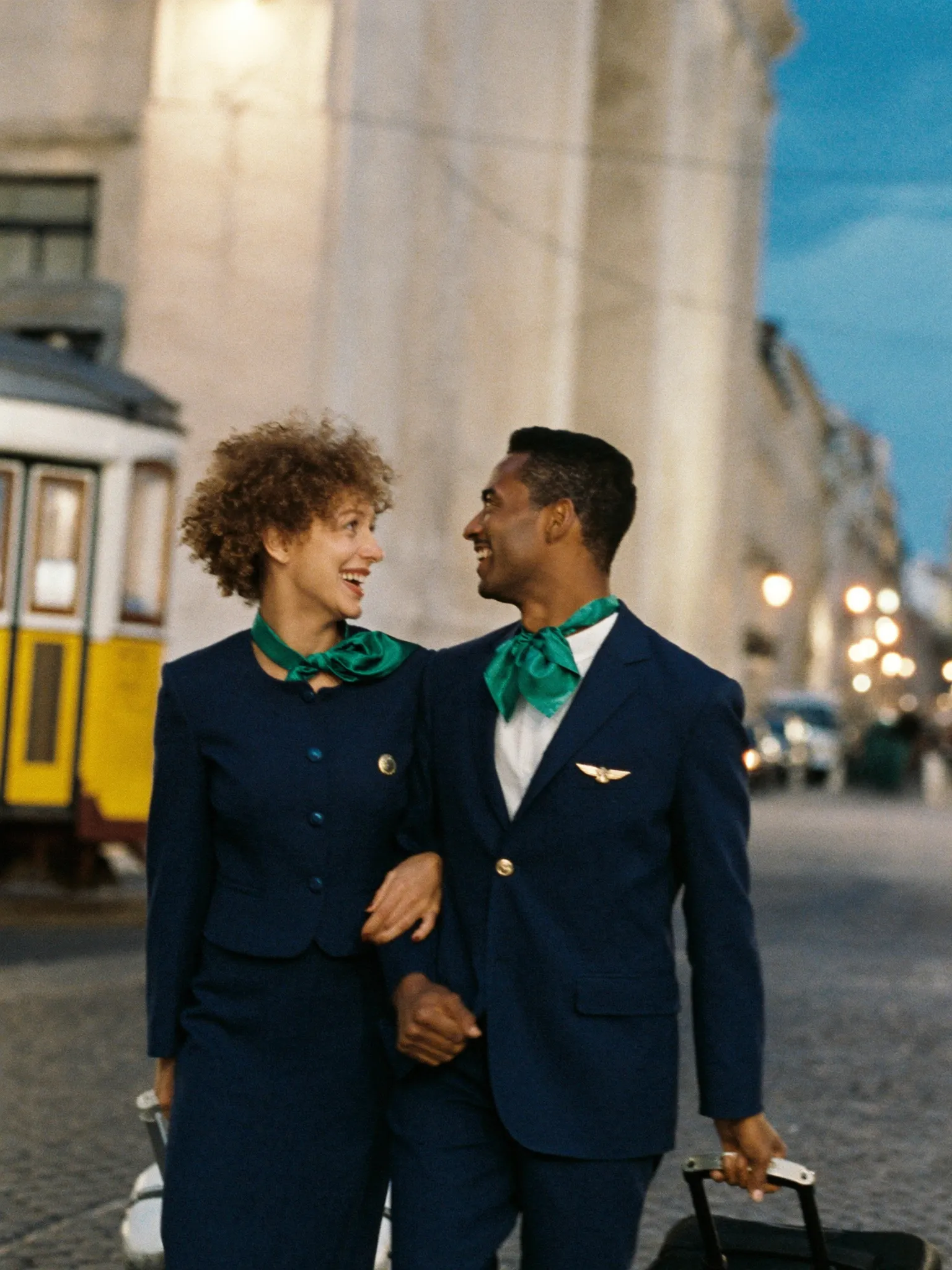 Dois comissários de voo rindo enquanto caminham com as malas por uma rua de Lisboa ao anoitecer